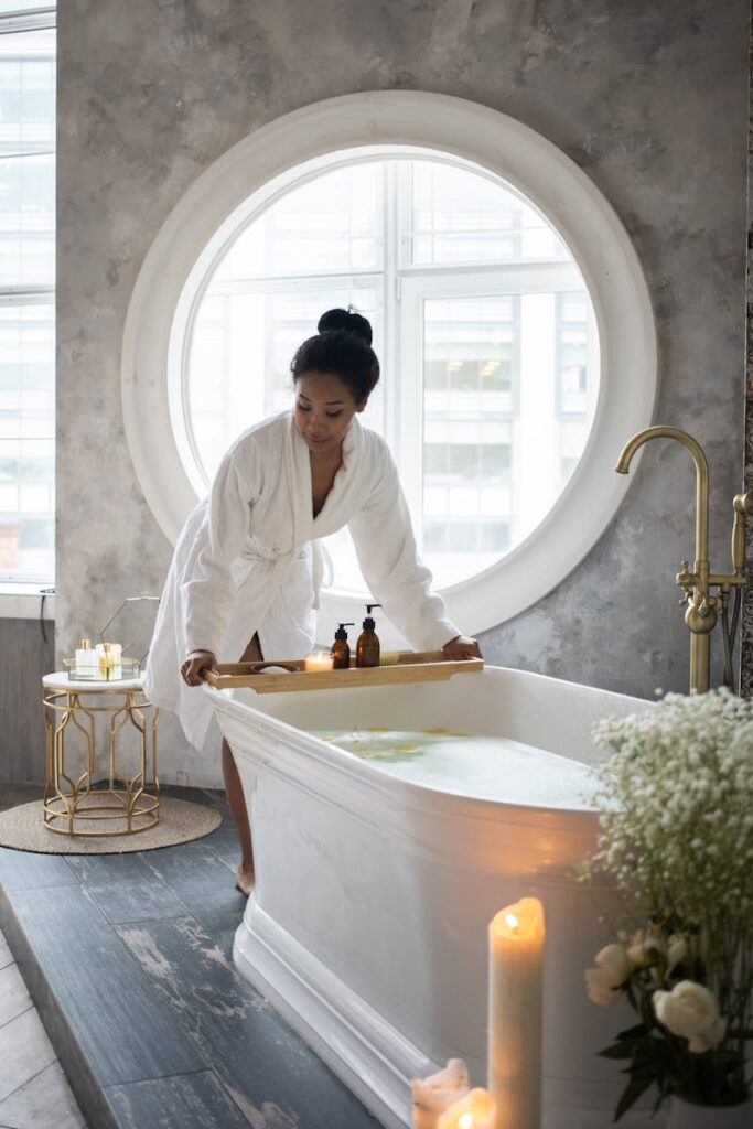 Woman in white robe preparing a spa bath with candles and flowers for relaxation.